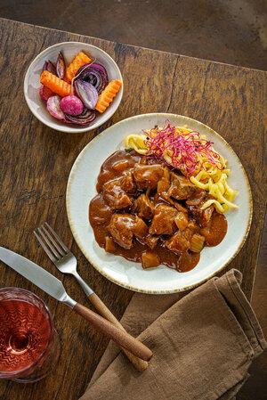 Goulash with spaetzle, garnished with red cress shoots and a selection of colourful roasted vegetables, served in a small bowl.